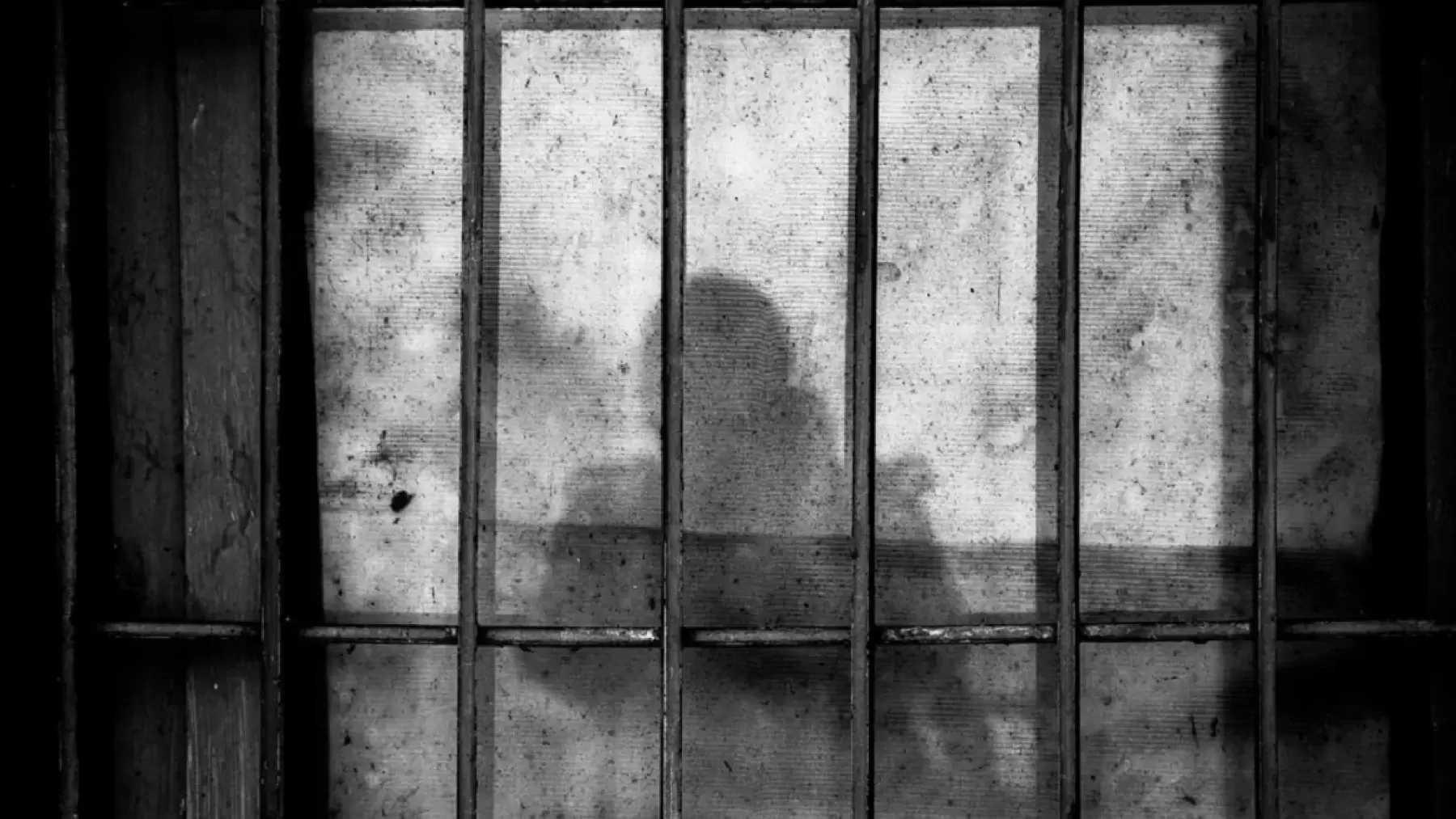 Image description: grey and black stock photo of bars on a window. A person's shadow is in the center of the window
